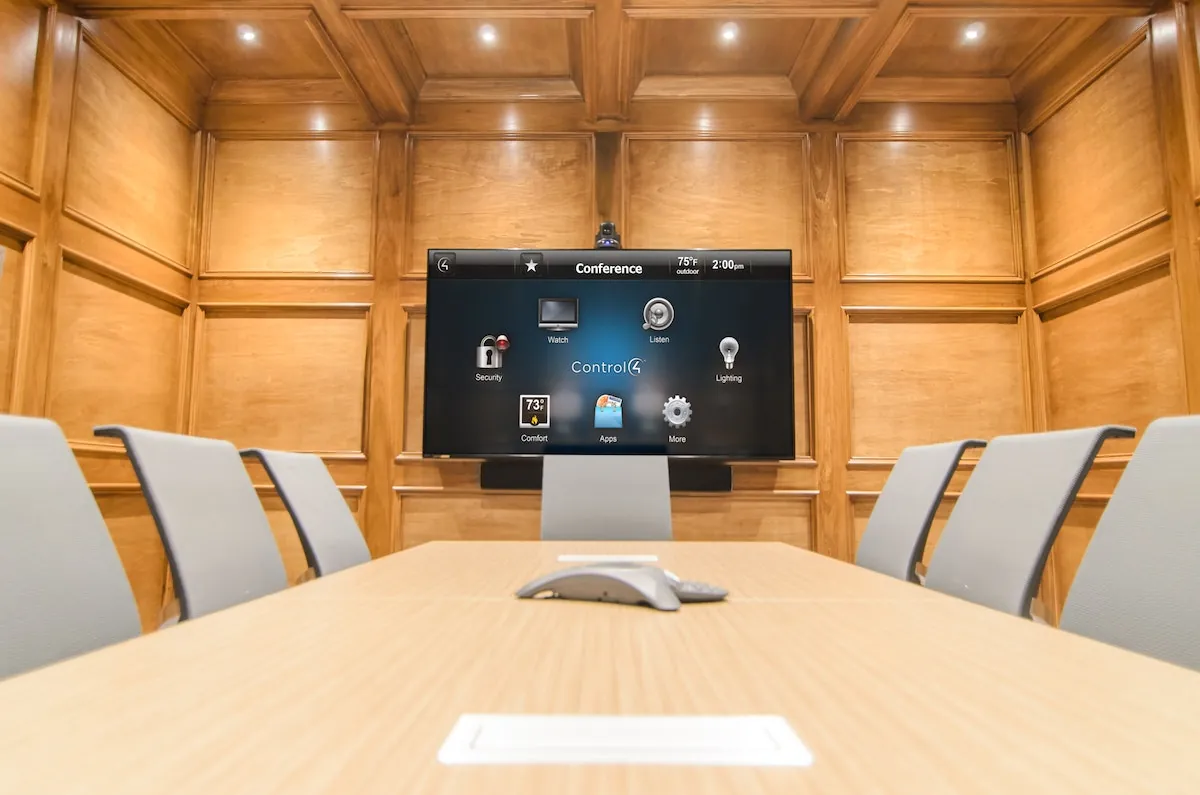 Wood-paneled conference room with a wall-mounted display showing a room control interface and a speakerphone on the table